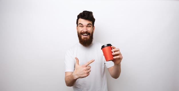 Bärtiger Mann, der eine rote Pappbecher Kaffee oder Tee hält und auf weißem Hintergrund im weißen T-Shirt darauf zeigt.