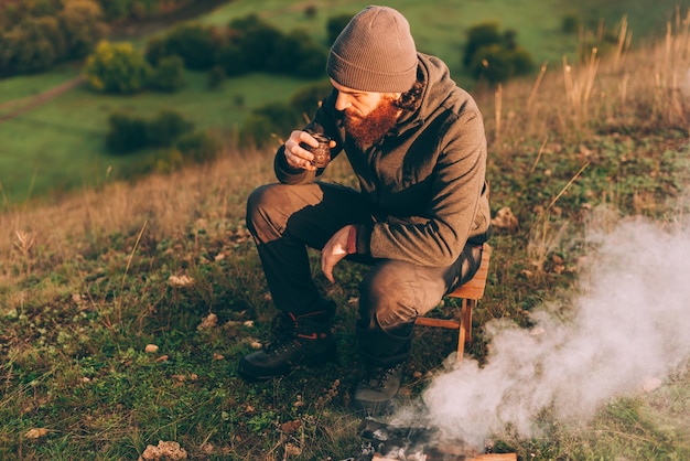 bärtiger Mann, der morgens Kaffee in der Nähe des Lagerfeuers trinkt