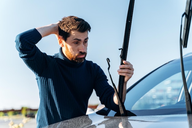 Bärtiger Mann, der sich verwirrt fühlt, während er während des sonnigen Tages die Scheibenwischer auf einem Auto auf der Straße wechselt Transportkonzept