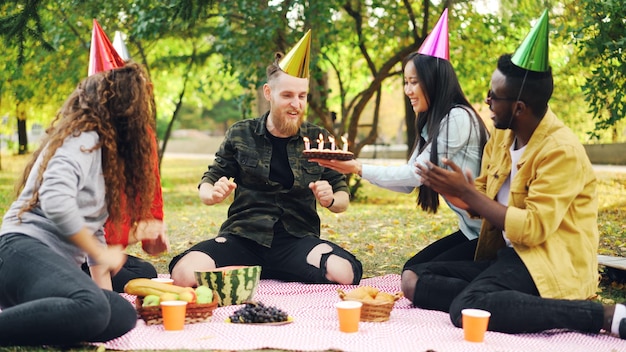 Bärtiger Mann feiert Geburtstag mit Freunden im Park, bläst Kerzen auf Kuchen, schreit und drückt positive Emotionen aus, während Freunde lachen und in die Hände klatschen