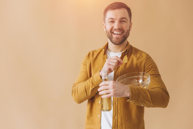 Foto bärtiger mann glücklich und lächelnd in gelbem hemd trinkt bier und isst chips auf beige hintergrund