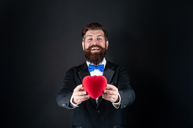 Bärtiger Mann im formellen Smoking mit Fliege teilen Liebesherz am 14. Februar Valentinstag, selektiver Fokus, Liebe.