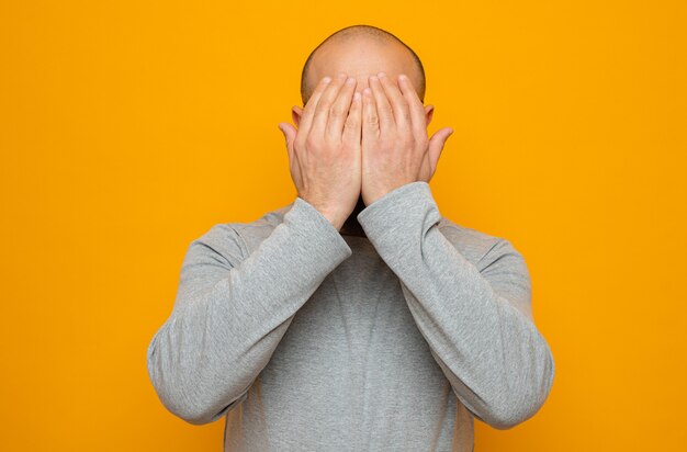 Foto bärtiger mann im grauen sweatshirt bedeckt die augen mit den händen, die über orangefarbenem hintergrund stehen