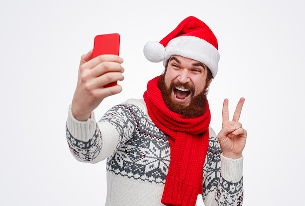 bärtiger Mann in der Weihnachtsmütze, der Friedenszeichen gestikuliert und selfie nimmt