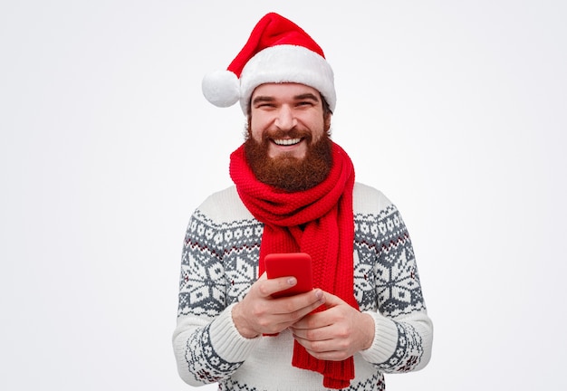 bärtiger Mann in der Weihnachtsmütze lächelnd beim Durchsuchen des Smartphones