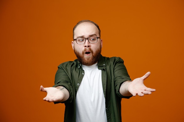 Foto bärtiger mann in freizeitkleidung mit brille, der in die kamera schaut, überrascht, wie er händchen vor ihm hält, als würde er etwas anbieten, das über orangefarbenem hintergrund steht