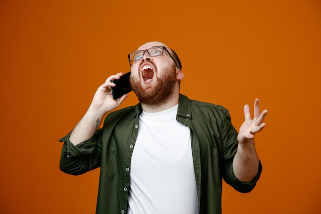Bärtiger mann in freizeitkleidung mit brille, der mit aggressivem gesichtsausdruck schreit, während er mit dem handy spricht, das über orangefarbenem hintergrund steht