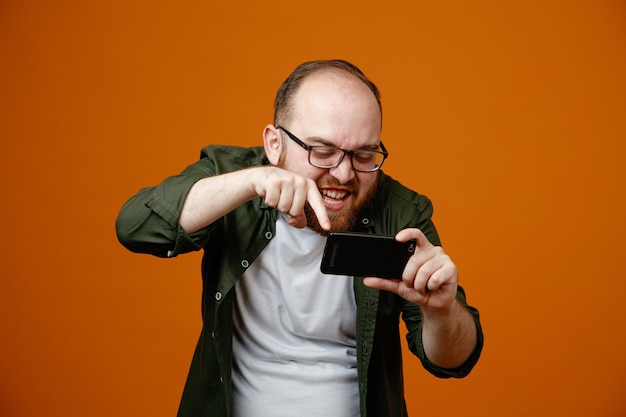 Bärtiger Mann in Freizeitkleidung mit Brille und Smartphone, der mit wütendem Gesicht und verärgertem Ausdruck auf orangefarbenem Hintergrund auf den Bildschirm blickt