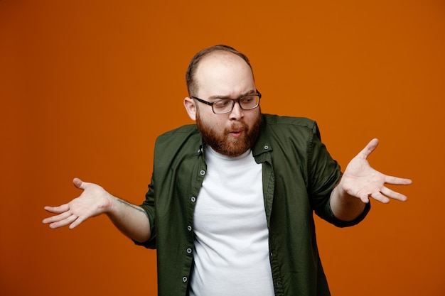 Bärtiger Mann in lässiger Kleidung mit Brille, der verwirrt aussieht und die Arme zu den Seiten ausbreitet, ohne eine Antwort auf orangefarbenem Hintergrund zu haben