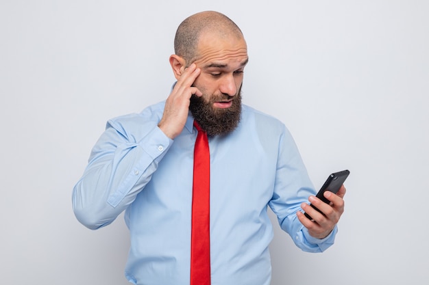 Bärtiger Mann in roter Krawatte und blauem Hemd, der das Handy hält und es erstaunt und überrascht anschaut