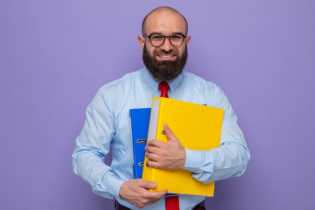Bärtiger Mann in roter Krawatte und blauem Hemd mit Brille, der Büroordner hält und in die Kamera schaut, die fröhlich auf lila Hintergrund steht