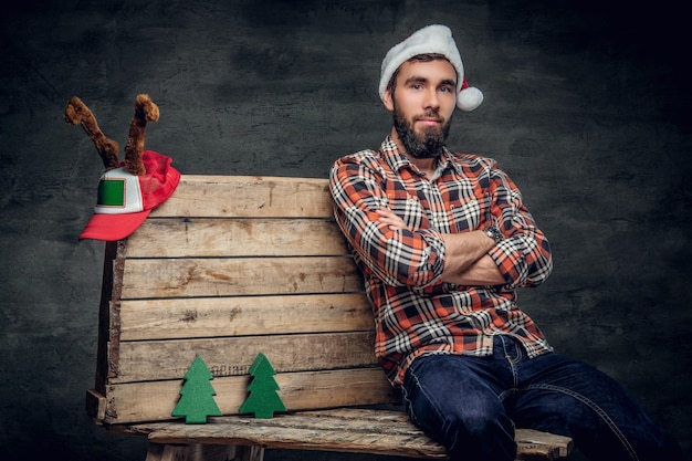 Bärtiger Mann in Santa's Hut und kariertem Hemd hält die Arme gekreuzt und sitzt auf einer Holzpalette mit kleinen geschmückten Tannen.