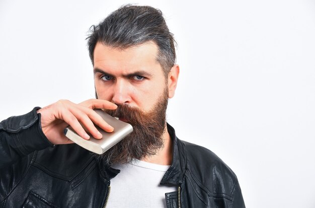 Bärtiger Mann, langer Bart, brutaler kaukasischer Hipster mit Schnurrbart, der metallische Flasche hält, Glas für alkoholisches Getränk, hat ernstes Gesicht, in schwarzer Lederjacke isoliert auf weißem Hintergrund, Kopierraum,