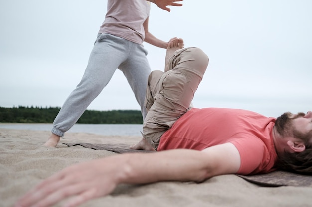 bärtiger mann macht yoga er macht übungen mit hilfe eines trainers, der auf einer matte an einem sandstrand liegt