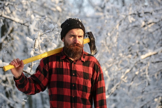 Bärtiger Mann mit Beil, Forstwirtschaft. Gut aussehender Mann, Hipster im verschneiten Wald. Holzfäller im Wald mit einer Axt am Wintertag. Männlich hält eine Axt auf einer Schulter. Brutaler bärtiger Mann. Männchen mit einer Axt.