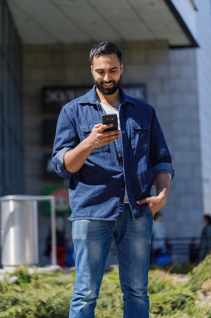 Bärtiger Mann mit einem Smartphone auf der Straße mit einem Bürogebäude im Hintergrund