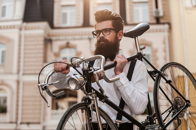 Bärtiger Mann mit Retro-Fahrrad. Niedriger Blickwinkel auf einen selbstbewussten jungen bärtigen Mann, der sein Fahrrad auf der Schulter trägt und beim Gehen im Freien wegschaut