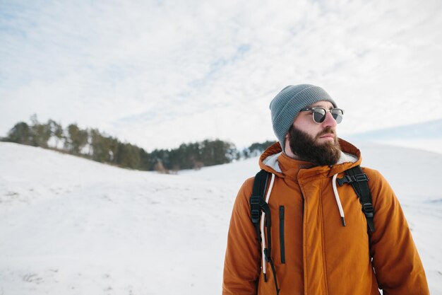 Bärtiger Mann mit Rucksack in Sonnenbrille und warmer Kleidung vor dem Hintergrund der Winterberge und des Waldes Das Konzept des Wanderns und des aktiven Lebensstils
