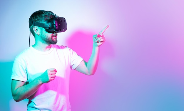 Bärtiger Mann mit VR-Brille, der mit dem Finger auf etwas zeigt, kommuniziert mit Freunden in einem Metaversum, Neon-Hintergrund, Augmented-Reality-Metaversum und Spielkonzept, virtueller Raum in der Zukunft