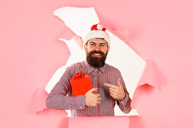 Foto bärtiger mann mit weihnachtsmannshut, der mit dem finger auf das weihnachtengeschenk zeigt, lächelnder mann in kariertem hemd, der sucht