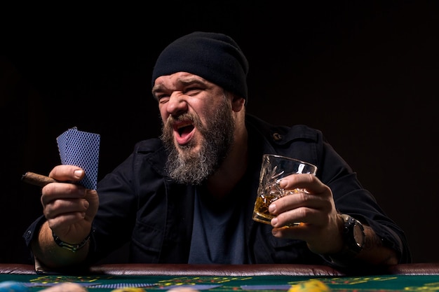Bärtiger Mann mit Zigarre und Glas sitzt am Pokertisch und schreit isoliert auf Schwarz