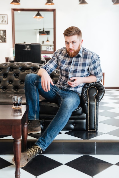 Bärtiger Mann sitzt auf dem Sofa im Friseursalon und schaut in die Kamera