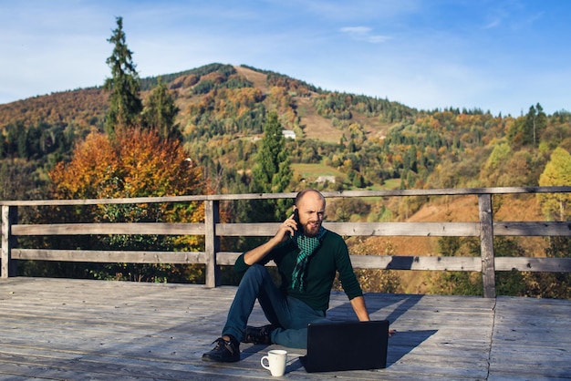 Bärtiger Mann sitzt mit Laptop auf der Terrasse in den Bergen und telefoniert.