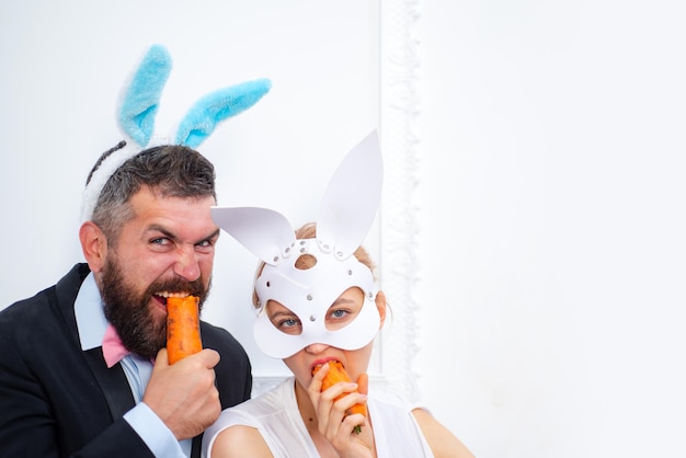 Bärtiger Mann und sexy Mädchen kopieren Raum Kaninchenfamilie mit Hasenohren Feiertagsfeiervorbereitung Osterhase frohe Ostern Frühlingsurlaub Eiersuche Osterhasenpaar isst orange Karotte