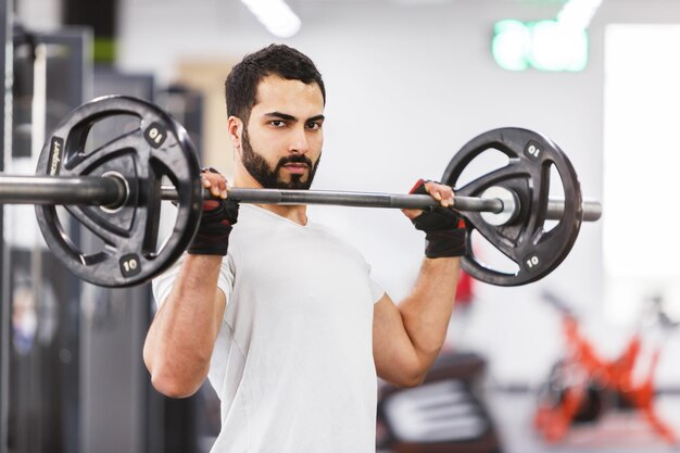 Bärtiger muskulöser Mann trägt weißes T-Shirt und trainiert mit Langhantel im Fitnessstudio