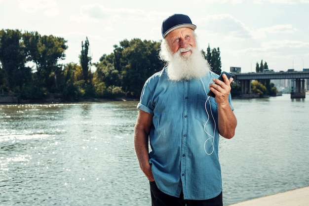 Bärtiger stilvoller reifer Mann in den Kopfhörern hört Musik unter Verwendung eines Smartphones. Glückliches Alter.