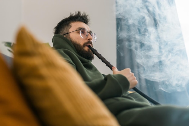 Bärtiger tausendjähriger oder gen z-mann, der shisha raucht, während er sich zu hause auf dem sofa entspannt und ausruht