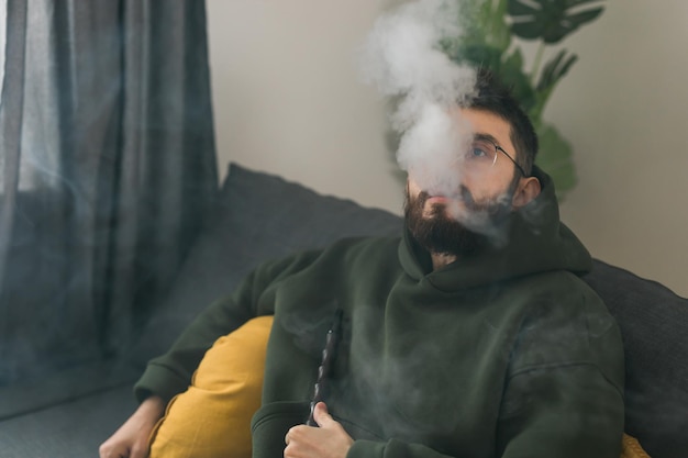 Bärtiger tausendjähriger oder gen z mann, der shisha raucht, während er sich zu hause auf dem sofa entspannt und ruhekonzept