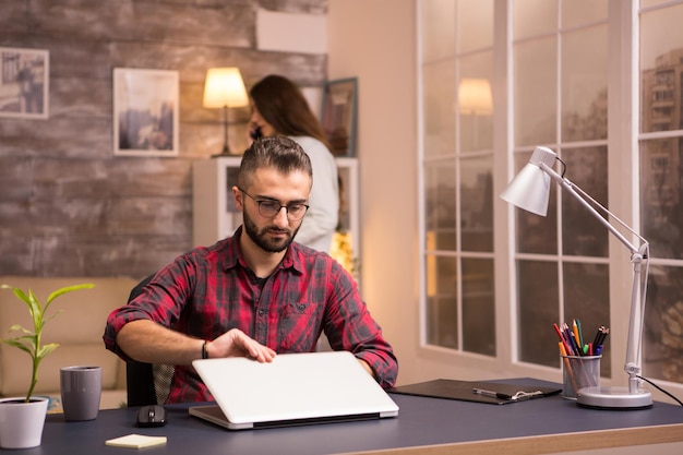 Foto bärtiger unternehmer öffnet laptop, um von zu hause aus zu arbeiten. freundin im hintergrund.