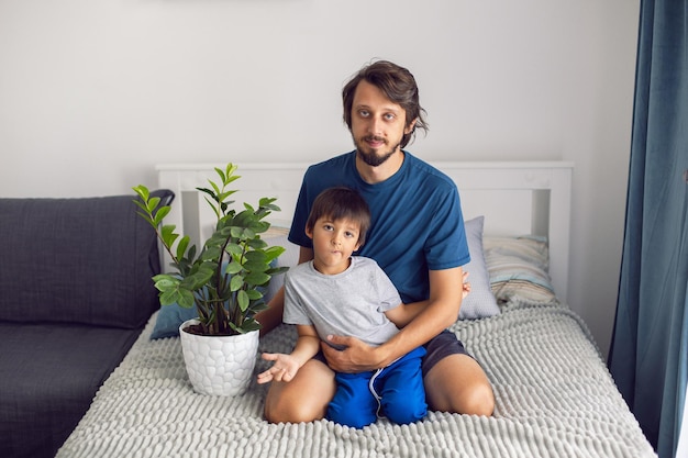 Bärtiger Vater und Sohn im T-Shirt sitzen auf einem Bett mit einer Blume in einem weißen Raum neben einem großen Fenster