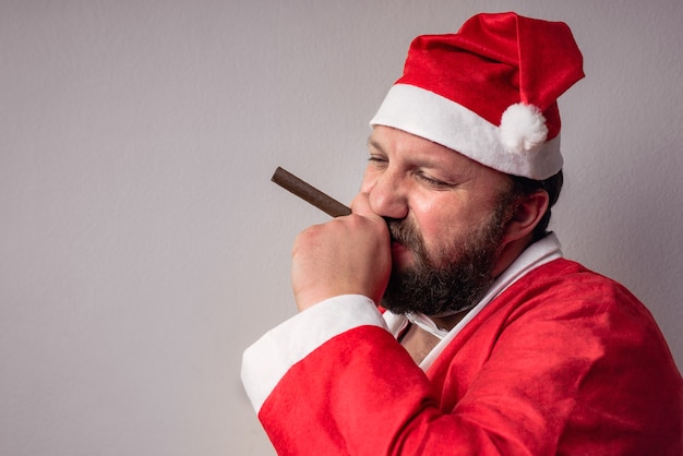 Bärtiger Weihnachtsmann mit langem Bart im Weihnachtspullover und Neujahrshut, der seine Zigarre raucht