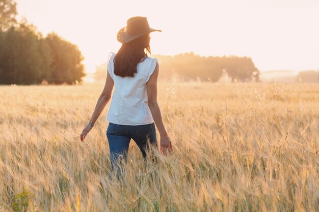 Bäuerin mit Cowboyhut, die bei Sonnenuntergang mit den Händen auf den Ohren auf dem landwirtschaftlichen Roggenfeld spazieren geht