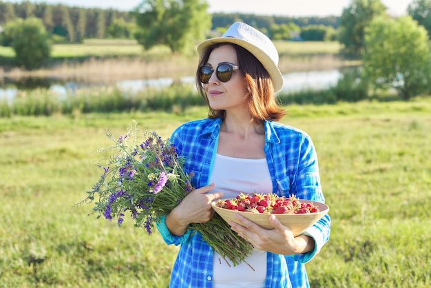 Bäuerin mit Schüssel frisch gepflückte Erdbeeren und Strauß Wildblumen. Naturhintergrund, ländliche Landschaft, grüne Wiese, Landhausstil