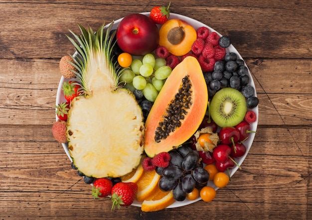 Bagas frescas de verão orgânico cru e frutas exóticas em chapa branca sobre fundo de madeira vintage. abacaxi, mamão, uva, nectarina, laranja, damasco, kiwi, morango, lichia, cereja. vista do topo