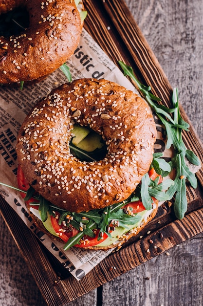 Bagels com creme, abacate, tomates e rúcula na placa de madeira.