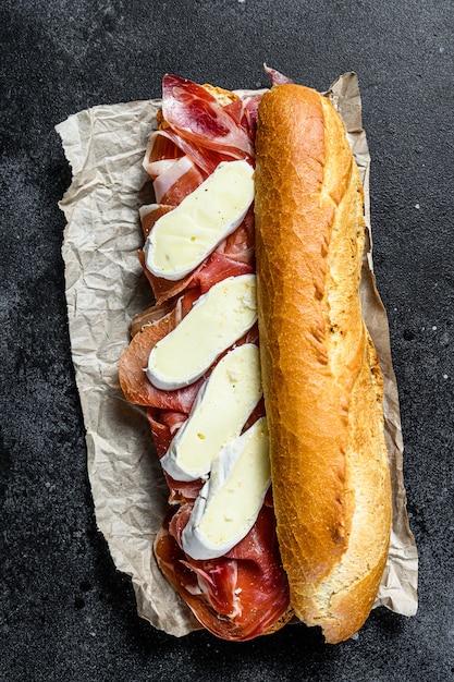 Baguette-Sandwich mit Jamon Ham Serrano, Paleta Iberica, Camembert-Käse. Schwarzer Hintergrund, Draufsicht.