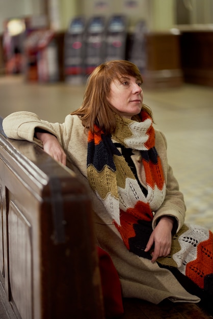 Bahnhof. Reife Frau in warmen Kleidern sitzt im Wartezimmer und wartet auf Zug. Der Abend ist dunkel. Vertikale