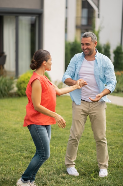 Bailando cerca de la casa. Esposa amorosa morena vestida con jeans bailando con su marido cerca de la casa de campo