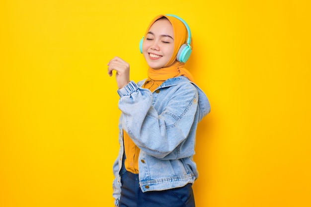 Bailando a una joven asiática con chaqueta de jeans usando audífonos para escuchar música aislada de fondo amarillo