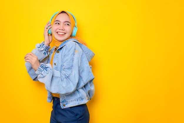 Bailando a una joven asiática con chaqueta de jeans usando audífonos para escuchar música aislada de fondo amarillo