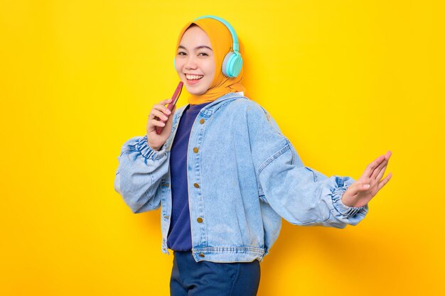 Bailando a una joven asiática con chaqueta de jeans usando audífonos para escuchar música y cantar a través de un teléfono móvil aislado en un fondo amarillo