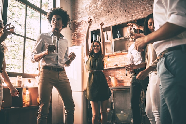 ¡Bailar es todo lo que necesitamos! Vista de ángulo bajo de jóvenes alegres bailando y bebiendo mientras disfruta de la fiesta en casa en la cocina