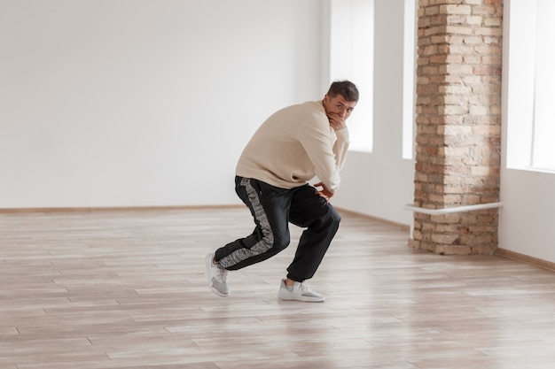 Bailarín de hip-hop joven y guapo bailando cubriendo su rostro con un suéter blanco y pantalón negro en el estudio de danza