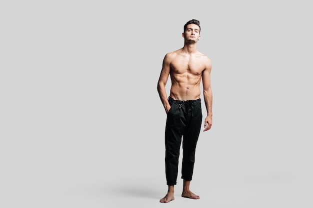 Foto bailarín joven guapo de pelo oscuro con el torso desnudo con un pantalón deportivo negro está de pie con la mano en los bolsillos sobre un fondo blanco