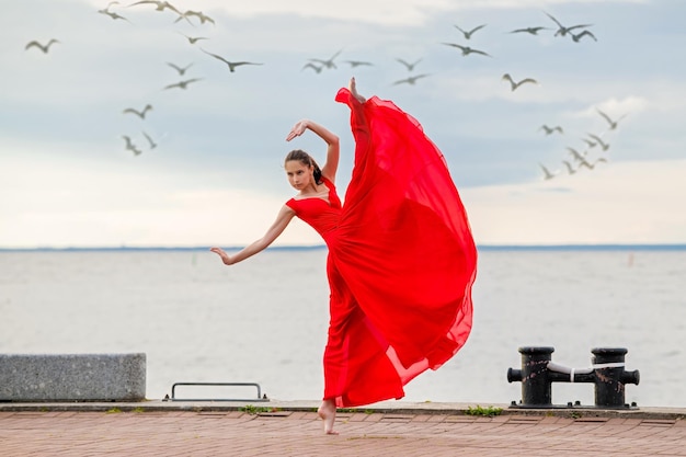 Bailarina bailarina con falda voladora roja y leotardo en el terraplén del océano o en la playa rodeada de gaviotas en el cielo