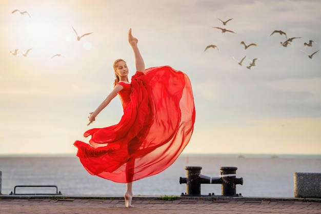 Bailarina bailarina con falda voladora roja y leotardo en el terraplén del océano o en la playa rodeada de gaviotas en el cielo